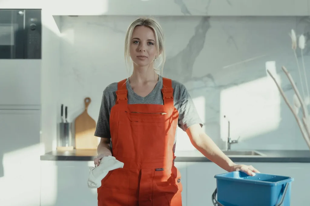 Domestic worker in red overalls standing in a modern UAE kitchen, holding a cleaning cloth and bucket